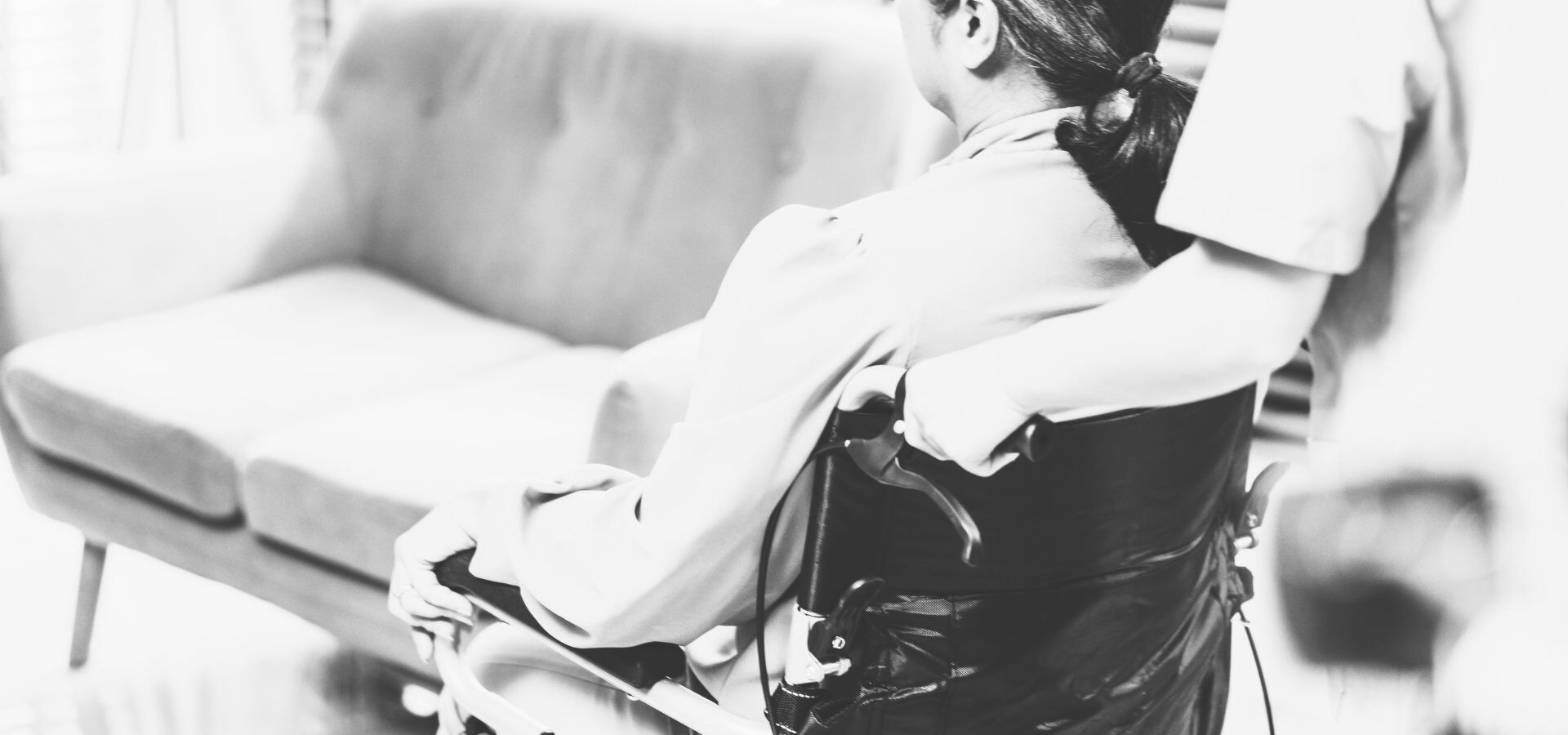 Eine Person im Rollstuhl wird von einem Pflegekraft durch einen Raum geschoben, im Hintergrund steht ein Sofa.